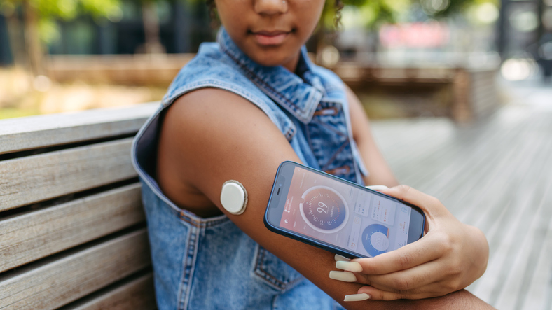 Diabetic woman sitting on bench in park, wearing a CGM sensor on her arm and checking her blood sugar level in real time with her smartphone.