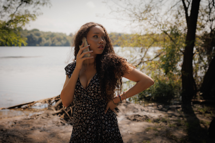 Curly-Haired Latin American Woman in a Forest Near a Lake While Using Her Mobile Phone on a Sunny Day