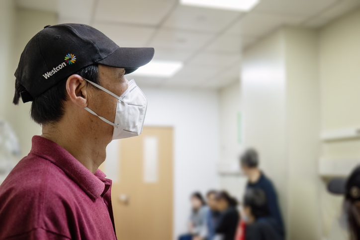 Man wearing a facemask, looking ahead absent minded. Unrecognizable people waiting in a corridor in the hospital.